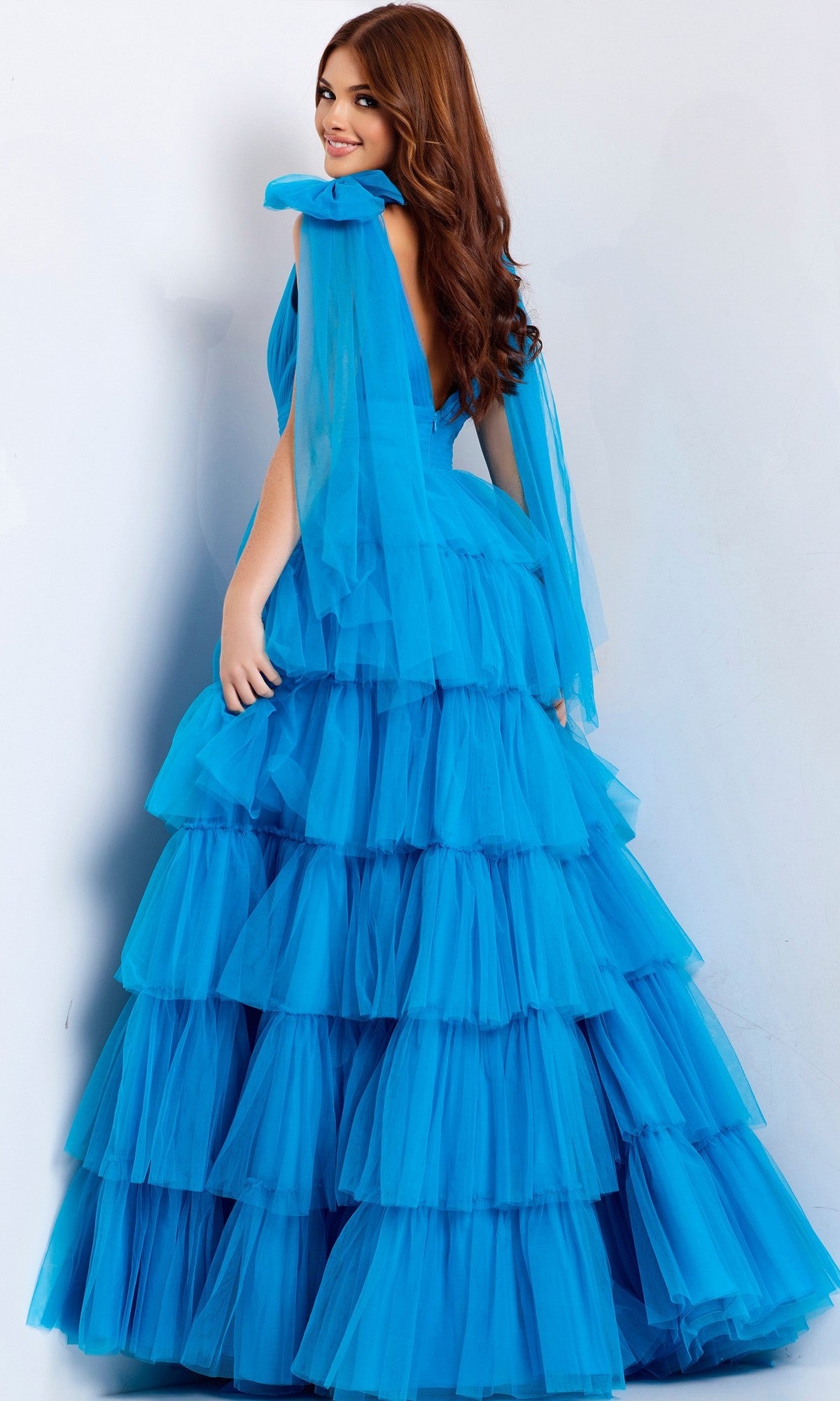 A woman with long brown hair models the Jovani 48288 long prom dress—a bright blue, sleeveless gown with a tiered ruffle skirt—while standing against a plain light background and glancing over her shoulder.